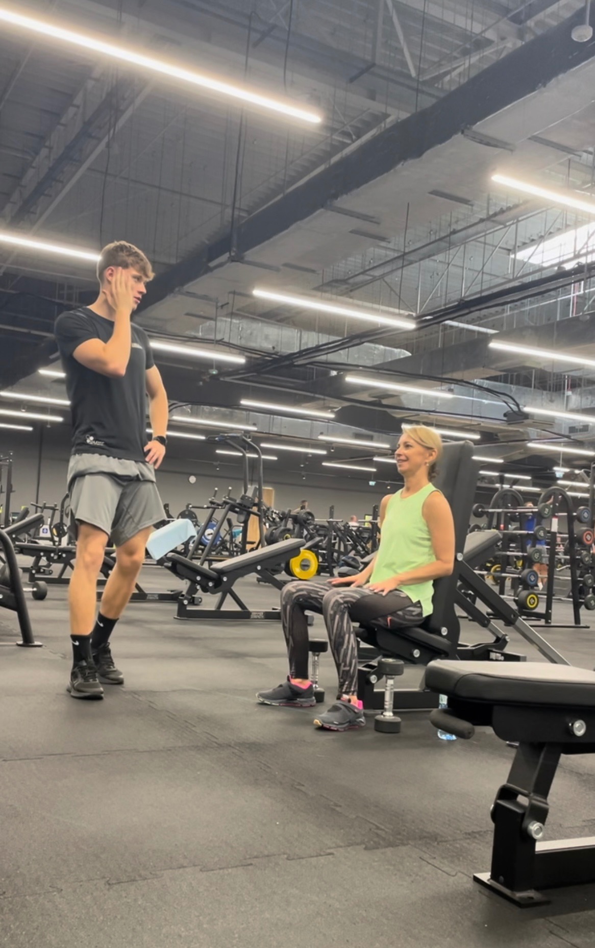 Kobieta siedzi na maszynie do ćwiczeń w siłowni, rozmawia z mężczyzną w sportowym stroju, który stoi obok z ręką na twarzy.