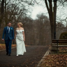 Rafał Torbicki Fotografia - Para młoda spaceruje alejką w parku, trzymając się za ręce. Pan młody w niebieskim garniturze, panna młoda w białej sukni z koronką. W tle drzewa i ławka.