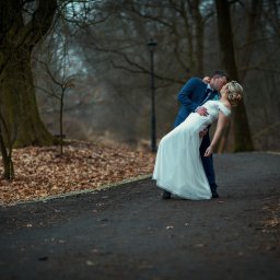 Rafał Torbicki Fotografia - Para młoda w jesiennej scenerii: Pan młody w niebieskim garniturze trzyma pannę młodą w białej sukni ślubnej, pochylając ją do pocałunku na tle drzew i opadłych liści.