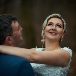Rafał Torbicki Fotografia - Uśmiechnięta panna młoda w objęciach pana młodego, spoglądająca w górę. Delikatny makijaż, blond włosy i biała suknia z koronki. Ujęcie w plenerze, z rozmytym tłem.