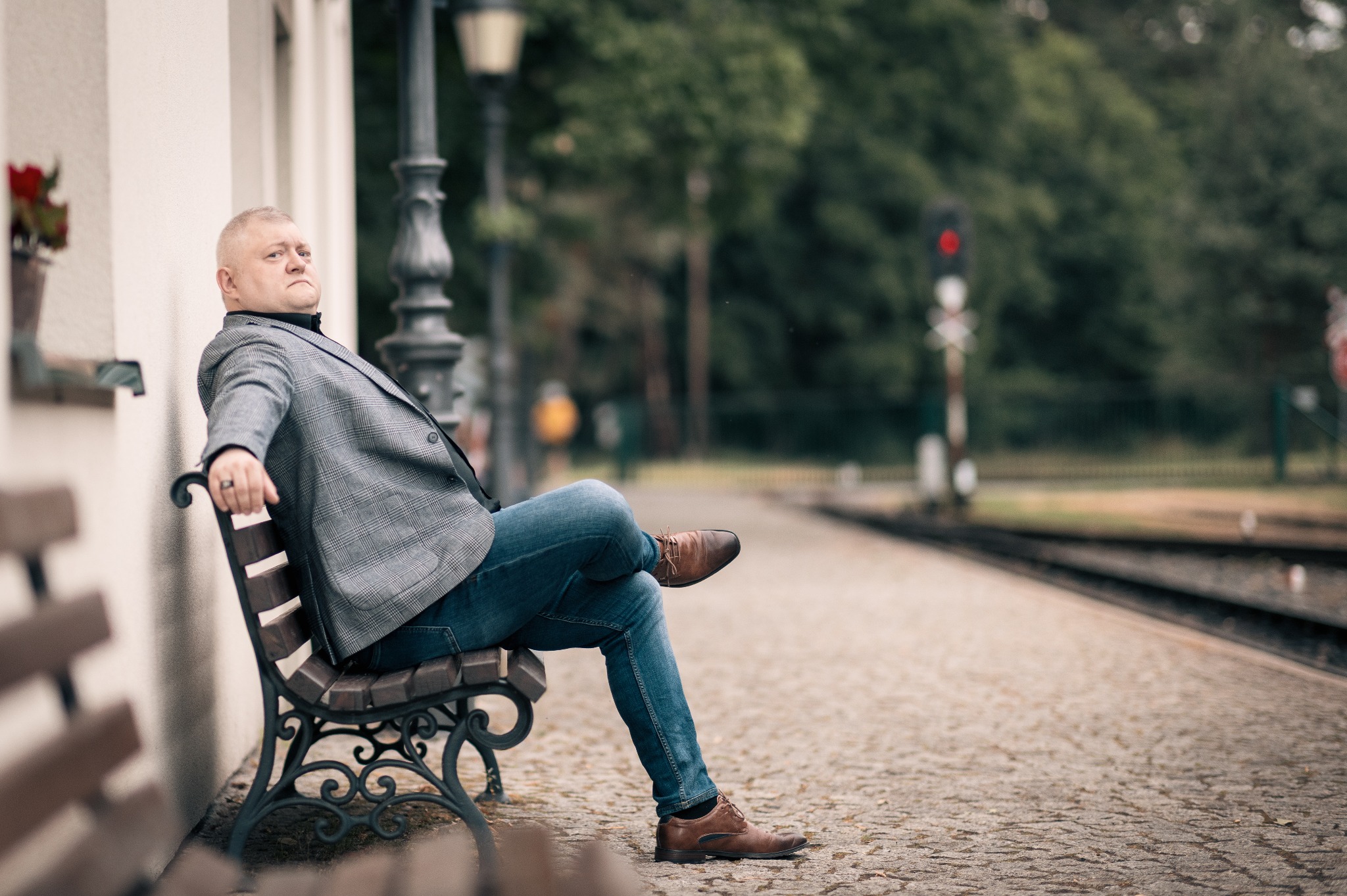 Mężczyzna w tweedowej marynarce relaksuje się na ławce peronowej, z nogą założoną na nogę, czekając na pociąg. W tle sygnalizacja kolejowa.