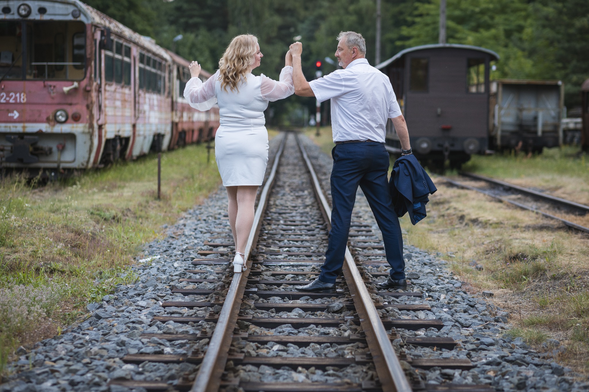 Para w jasnych strojach spaceruje po torach kolejowych, trzymając się za ręce; w tle stare wagony i zieleń. Nietypowa sesja w plenerze.