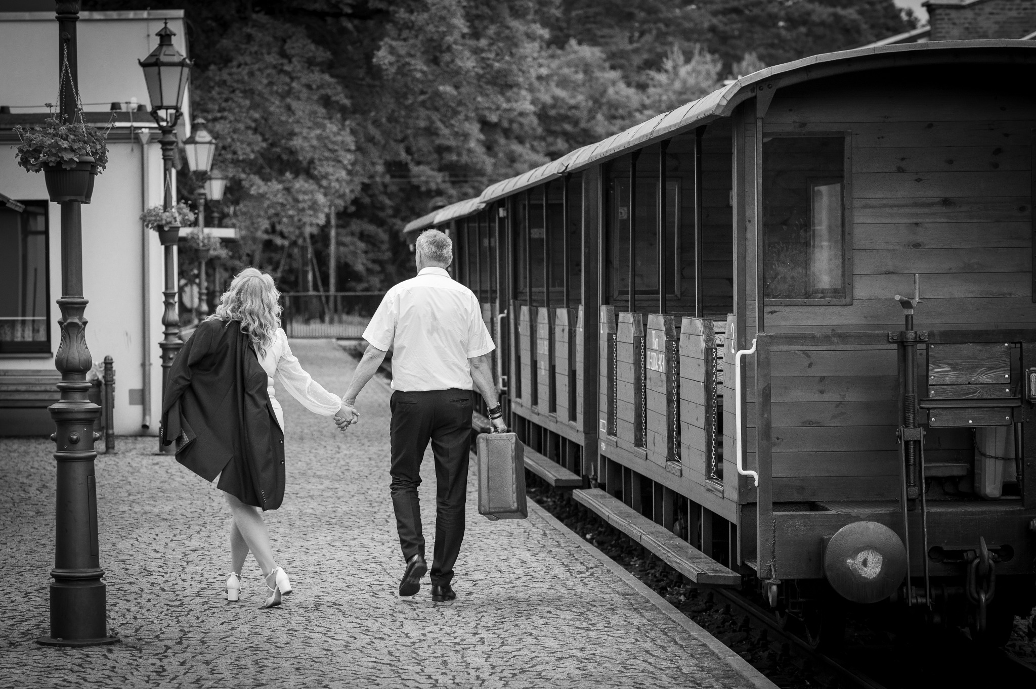 Czarno-białe zdjęcie pary trzymającej się za ręce na peronie, obok wagonu kolejowego. Kobieta w jasnej sukience i marynarce, mężczyzna z walizką. Romantyczna atmosfera.