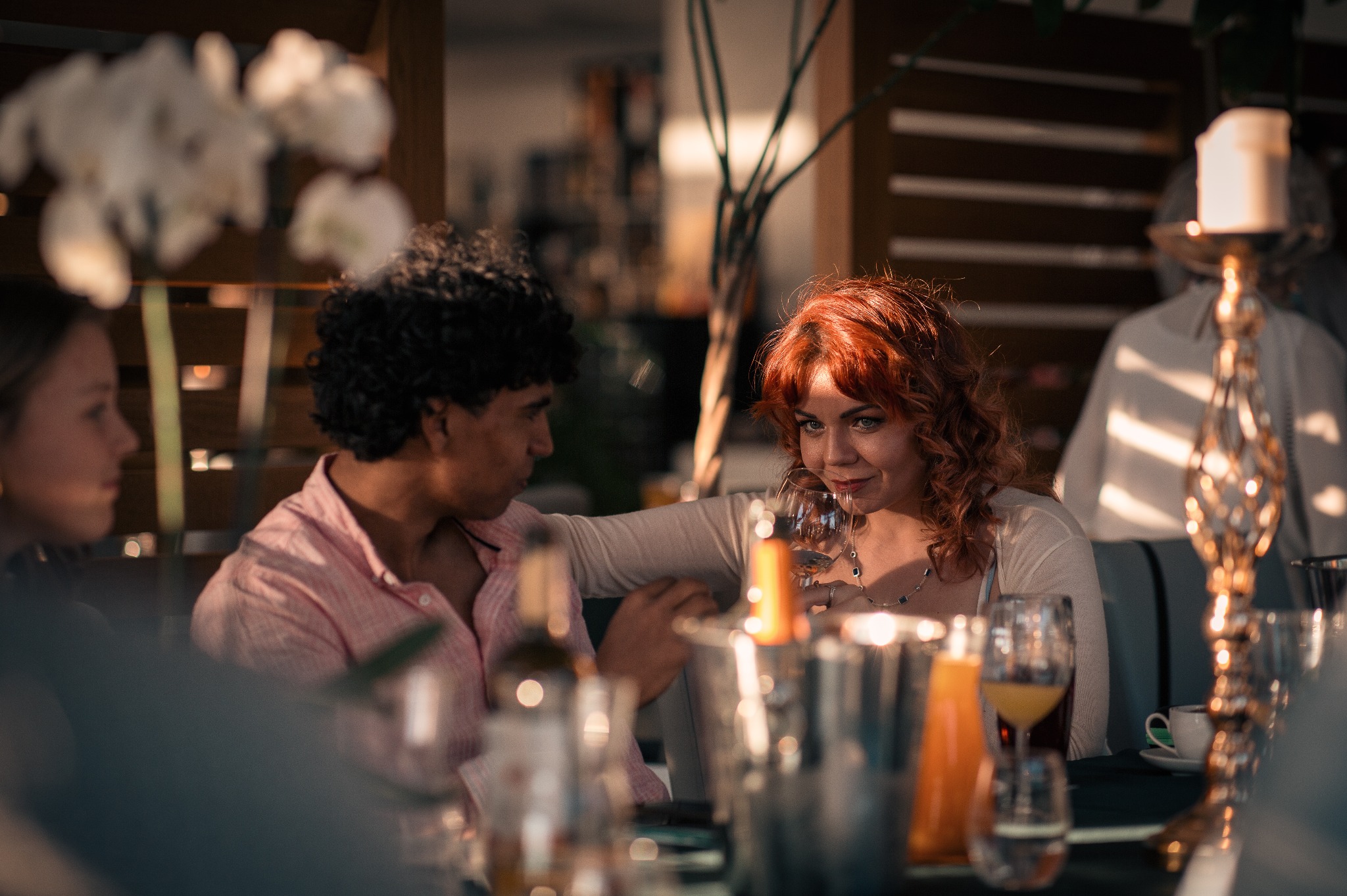 Elegancka para siedzi przy stole z napojami, kobieta z rudymi włosami trzyma kieliszek wina, delikatne światło podkreśla intymną atmosferę spotkania.