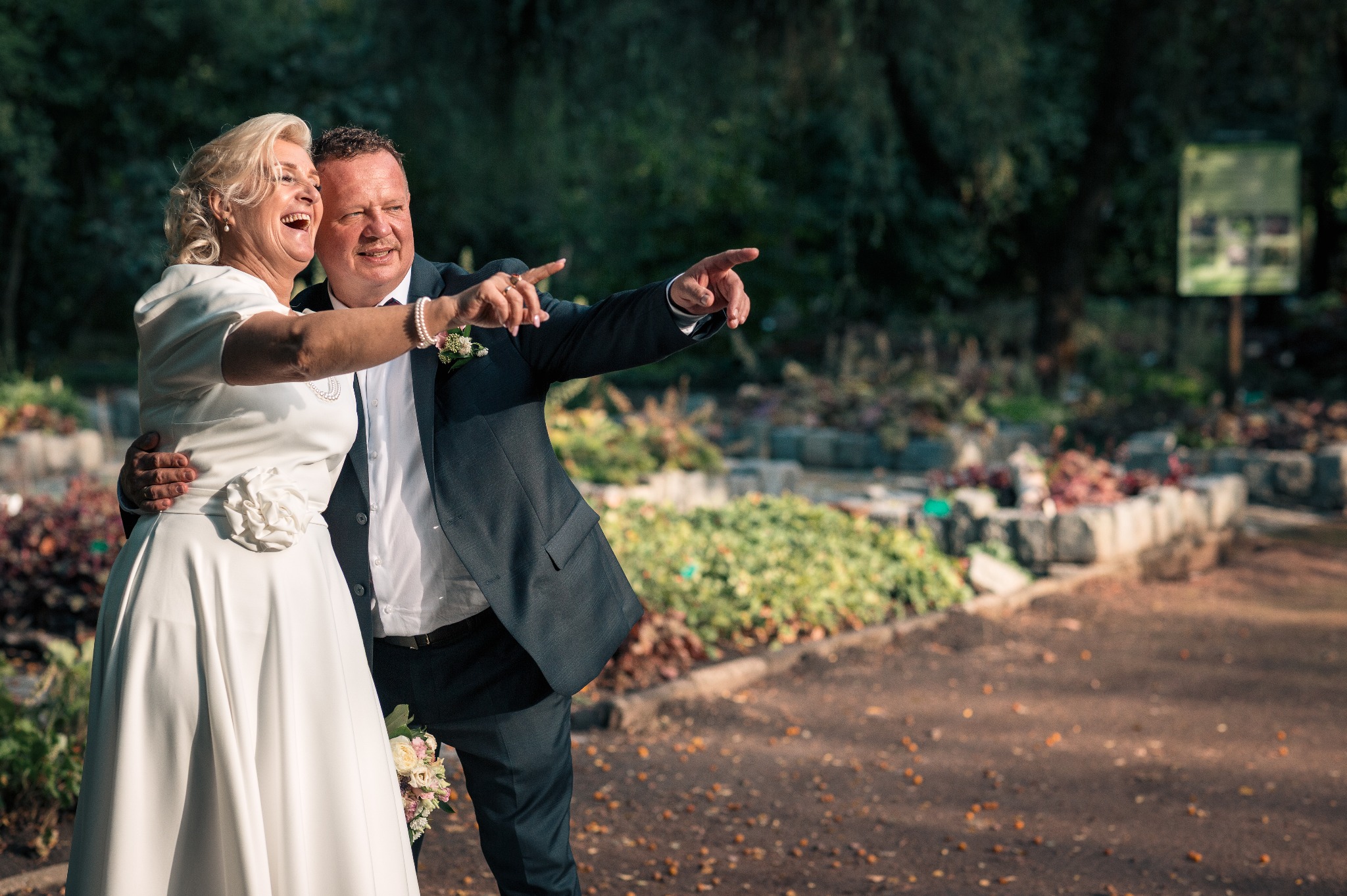 Uśmiechnięta para w strojach ślubnych, obejmująca się w parku, wskazująca palcami w dal. Ona w białej sukni, on w ciemnym garniturze. W tle zieleń i ścieżka.