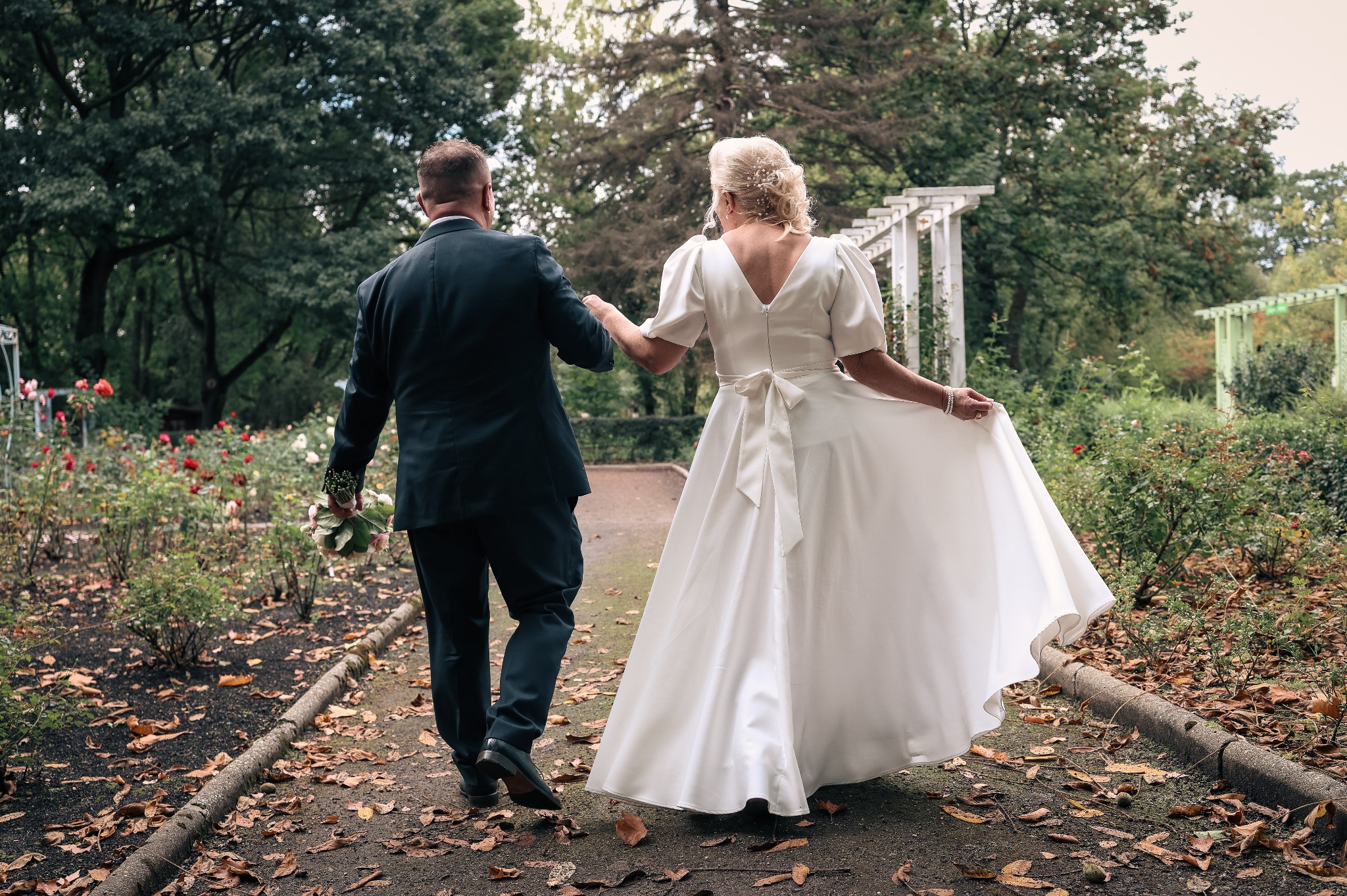 Para młoda idzie ścieżką w parku, trzymając się za ręce. Panna młoda w białej sukni z kokardą, pan młody w ciemnym garniturze. Jesienne liście na ziemi.