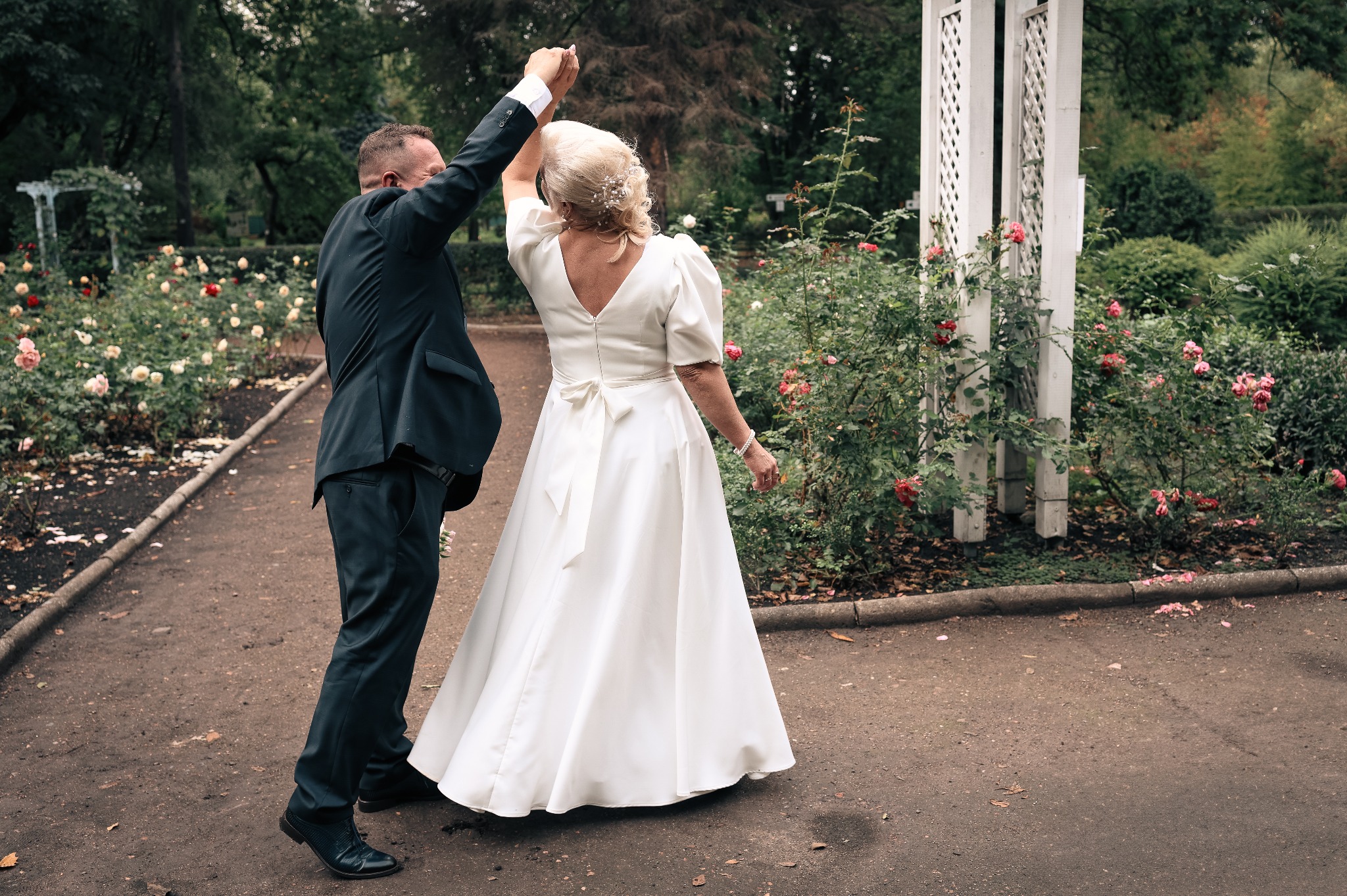 Para młoda tańczy w ogrodzie różanym, trzymając się za ręce. Panna młoda w białej sukni, pan młody w ciemnym garniturze. W tle biała altana i bujna roślinność.