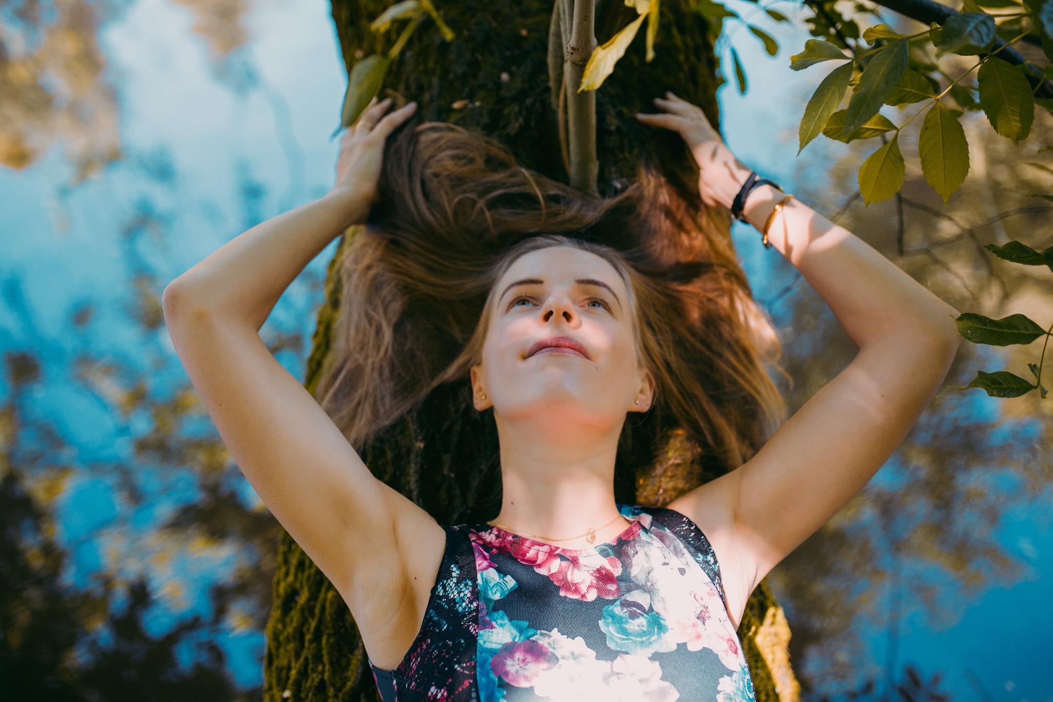 Młoda kobieta w kwiecistej sukience leży u podnóża drzewa, ręce uniesione nad głową, spoglądająca w górę na tle błękitnego nieba i zielonych liści.