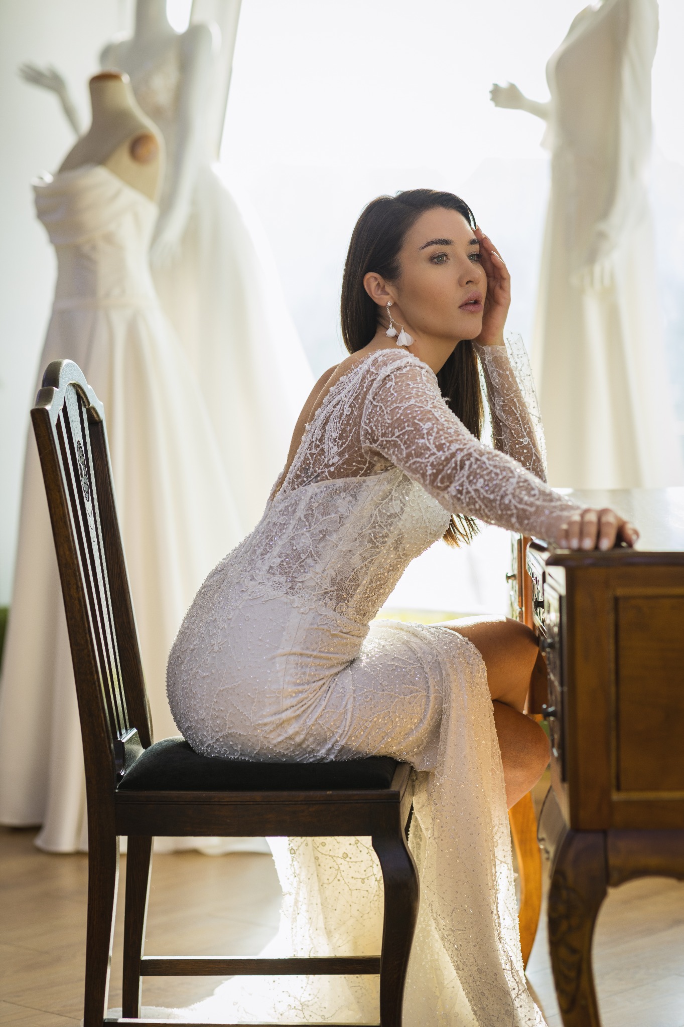 Elegancka brunetka w koronkowej sukni ślubnej siedzi przy drewnianym biurku. W tle manekiny z sukniami ślubnymi i jasne okno.