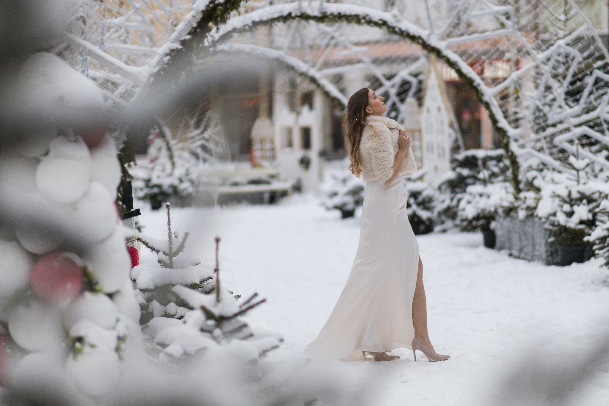 Kobieta w białej sukni i futerku pozuje w zimowej scenerii, z łukami pokrytymi śniegiem w tle. Ujęcie z dołu podkreśla elegancję modelki i magiczny klimat otoczenia.
