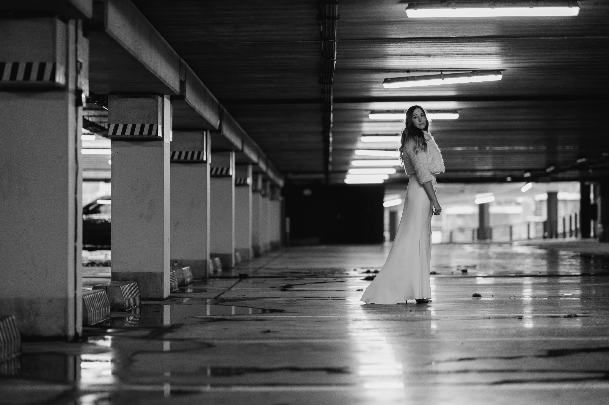Czarno-białe zdjęcie kobiety w sukni wieczorowej na parkingu podziemnym. Nietypowa lokalizacja, surowy klimat, oświetlenie jarzeniowe. Ujęcie z dołu, podkreślające sylwetkę.