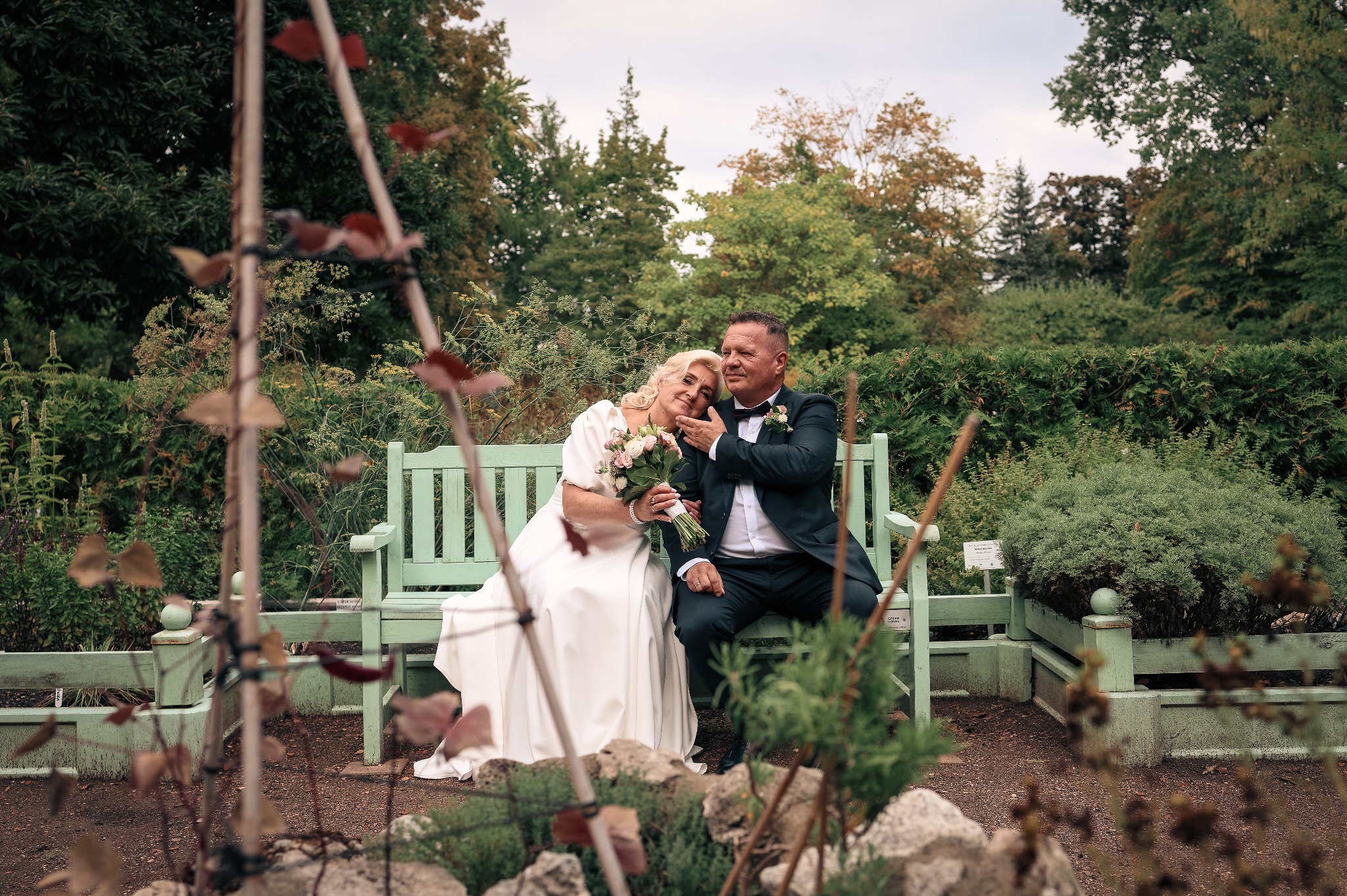 Para w strojach ślubnych siedzi na ławce w otoczeniu zieleni. Panna młoda trzyma bukiet kwiatów, a pan młody obejmuje ją czule. Romantyczna atmosfera w plenerze.