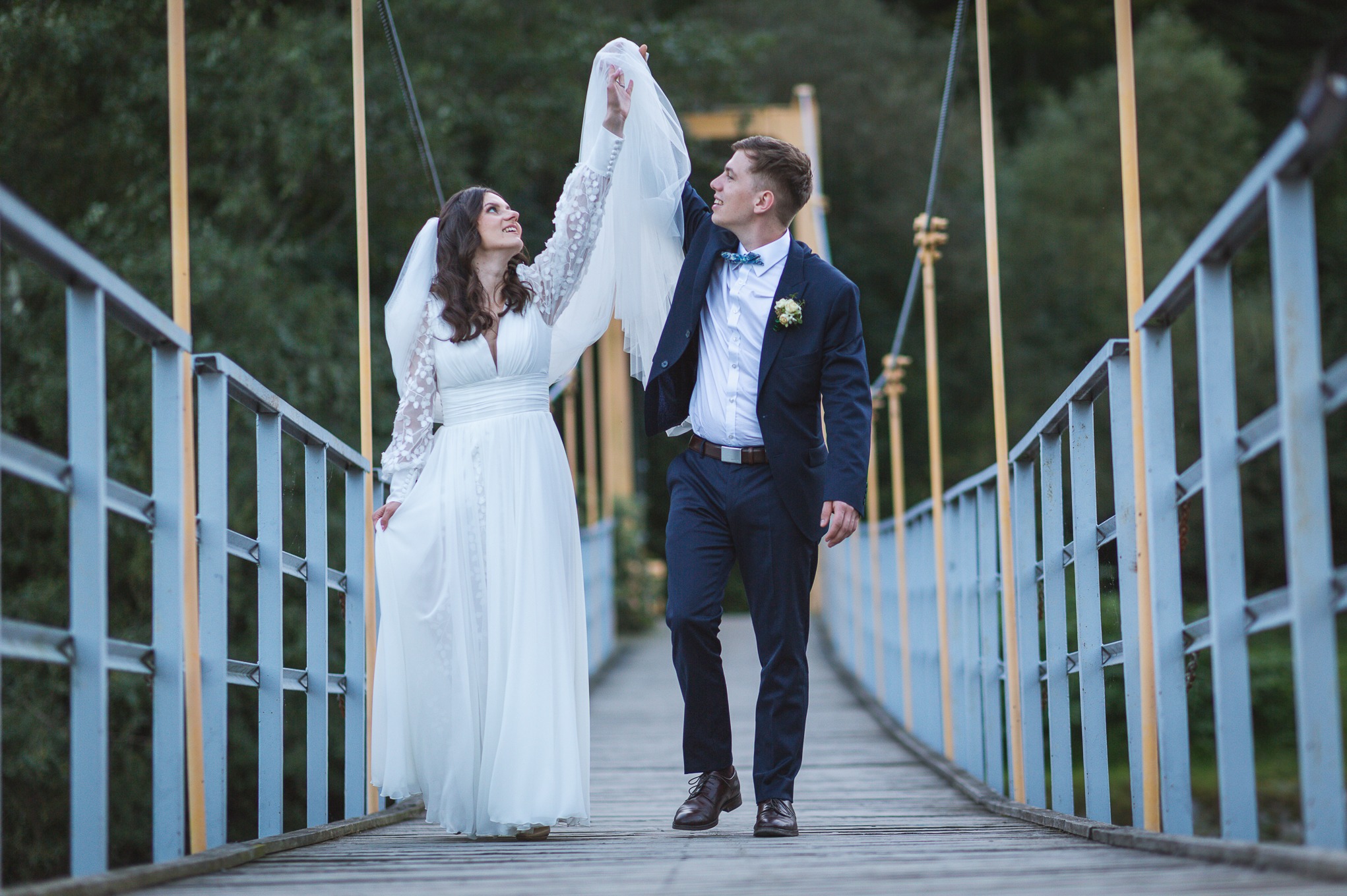 Para młoda na moście: panna młoda w sukni z koronkowymi rękawami trzyma welon, a pan młody w garniturze z muchą, oboje patrzą w górę.