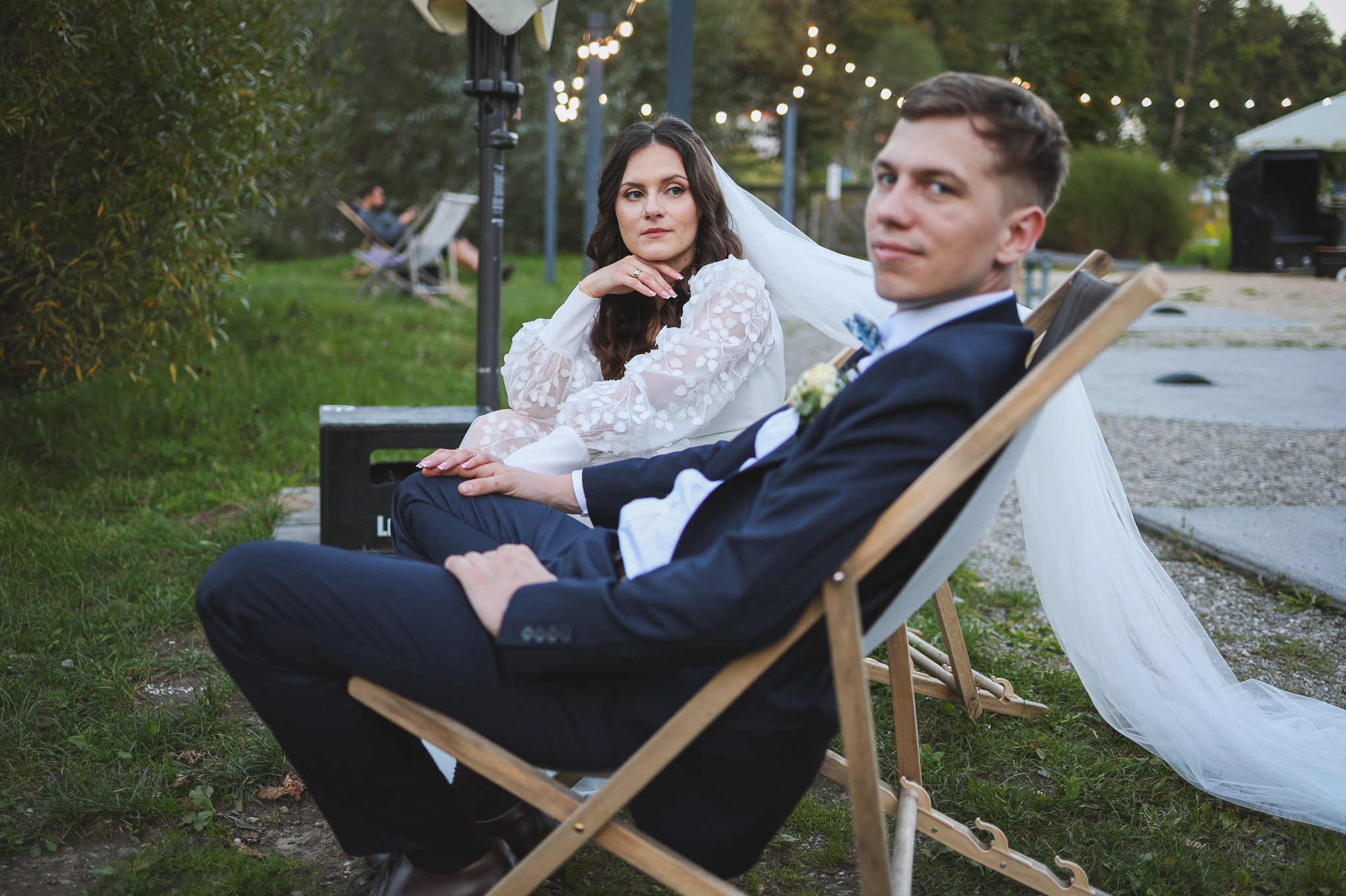 Para młoda siedzi na leżakach w plenerze. Ona w sukni z koronkowymi rękawami, on w granatowym garniturze. W tle drzewa i girlandy żarówek.