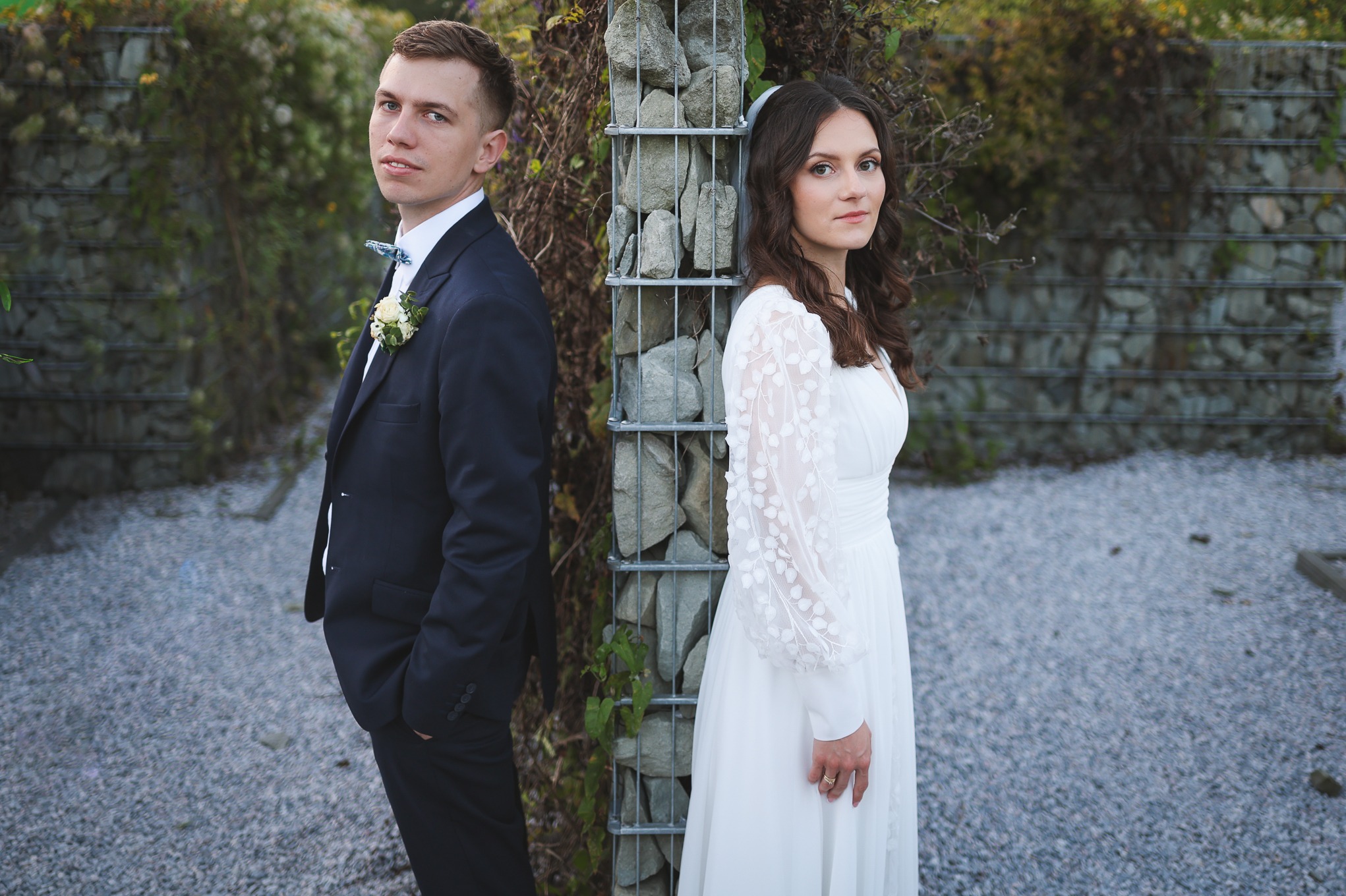 Para młoda w Rydułtowach pozuje na tle kamiennej ściany porośniętej zielenią. Pan młody w ciemnym garniturze, panna młoda w białej sukni z długim rękawem.
