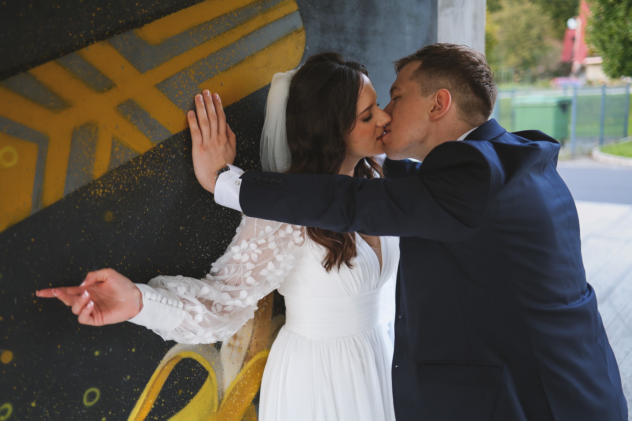 Pocałunek młodej pary na tle graffiti: Panna Młoda w zwiewnej sukni z koronkowymi rękawami opiera dłoń o ścianę, Pan Młody w granatowym garniturze obejmuje ukochaną.