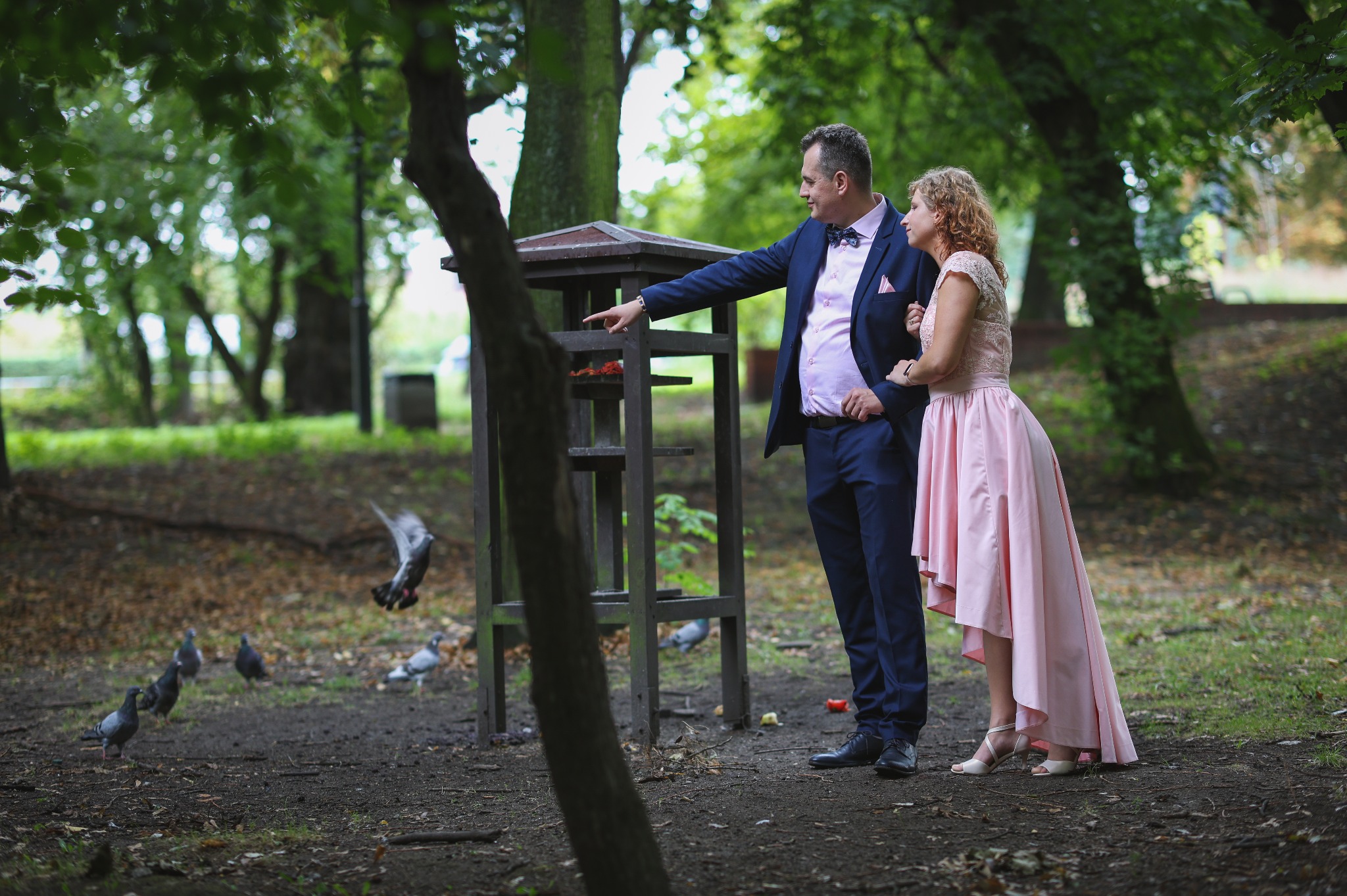 Elegancka para w parku, mężczyzna w granatowym garniturze wskazuje na karmnik dla ptaków, kobieta w różowej sukience, gołębie w tle, naturalne światło.