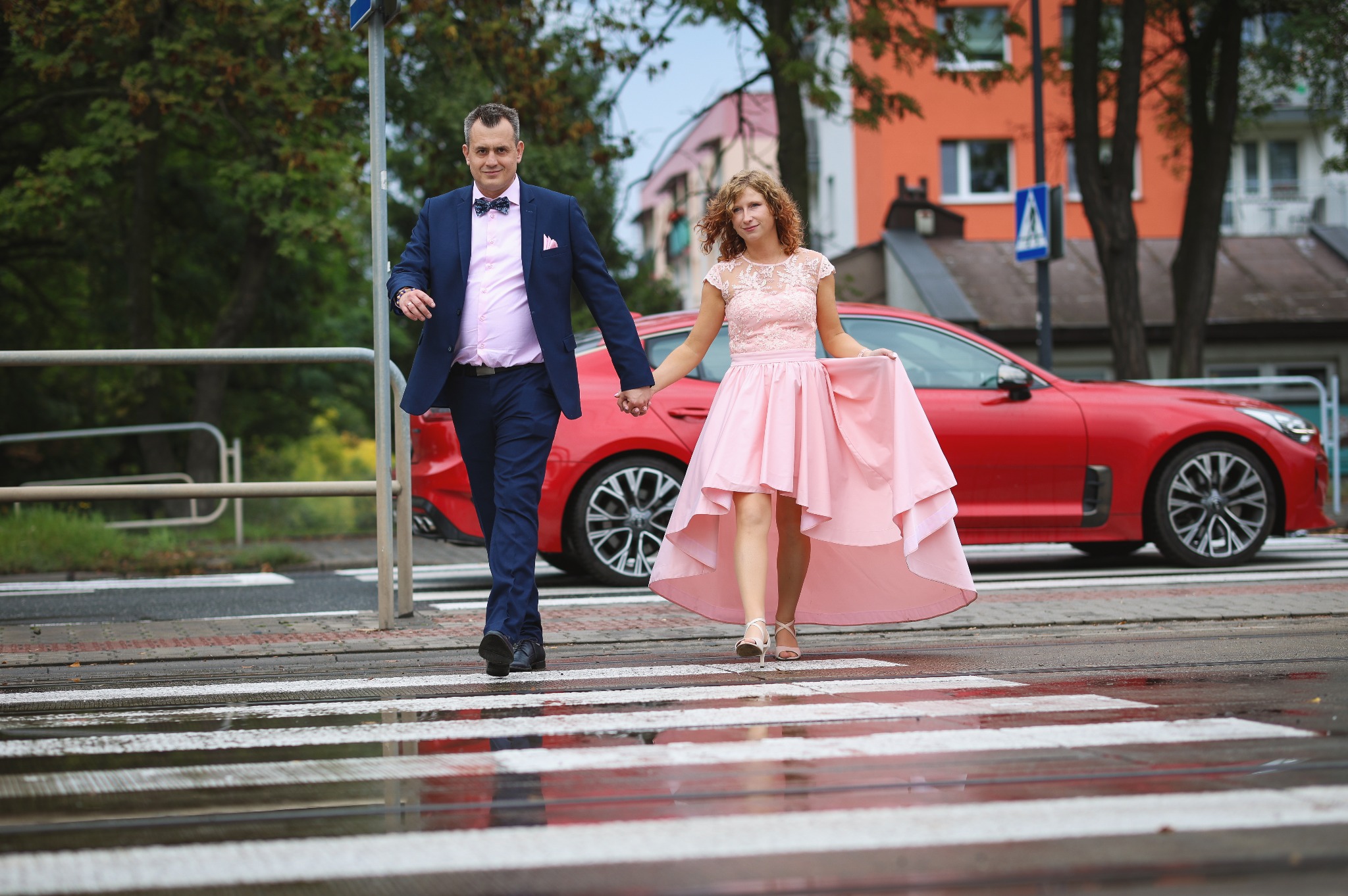 Para w eleganckich strojach, on w granatowym garniturze i muszce, ona w różowej sukience, trzymają się za ręce na mokrym przejściu dla pieszych, z czerwonym autem w tle.