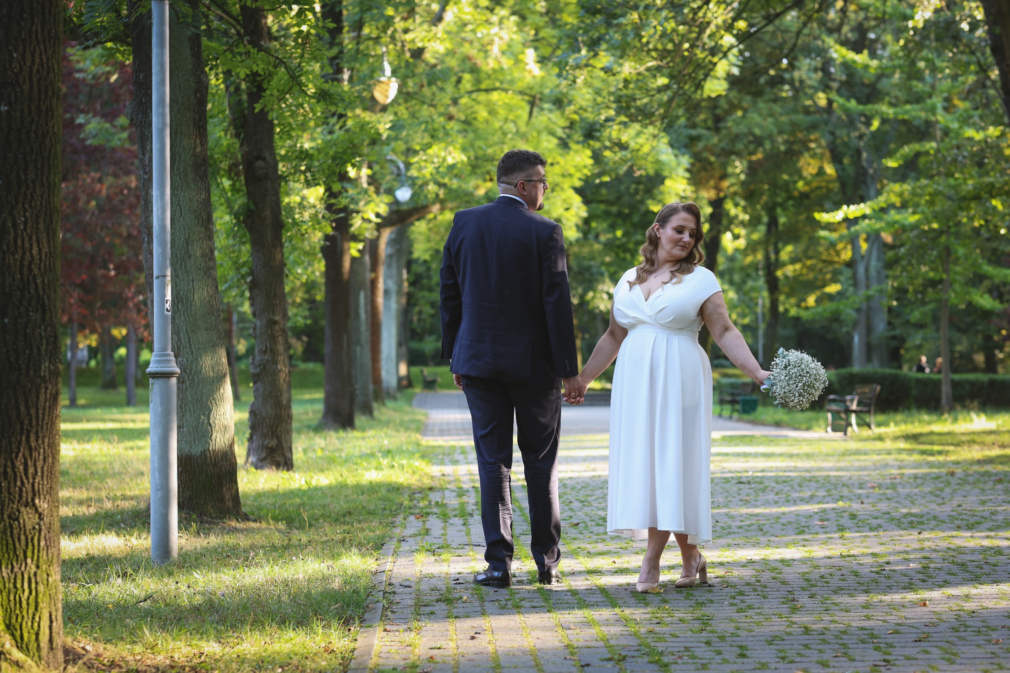 Para młoda spaceruje po parku, trzymając się za ręce. Panna młoda w białej sukni, pan młody w garniturze. Ujęcie z tyłu, z widoczną alejką i drzewami w tle.