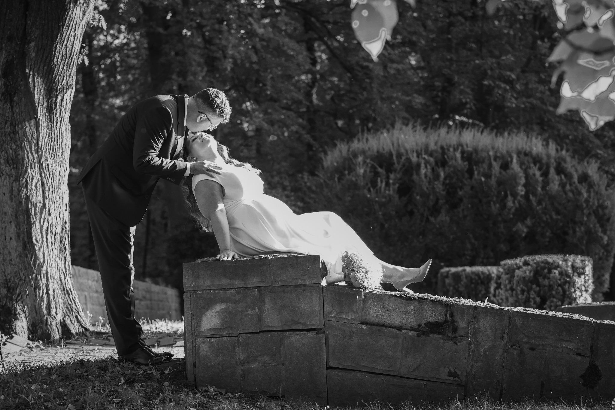 Czarno-białe zdjęcie pary młodej: pan młody w garniturze całuje pannę młodą w sukni, która leży na kamiennym murku w parku. W tle drzewa i krzewy.
