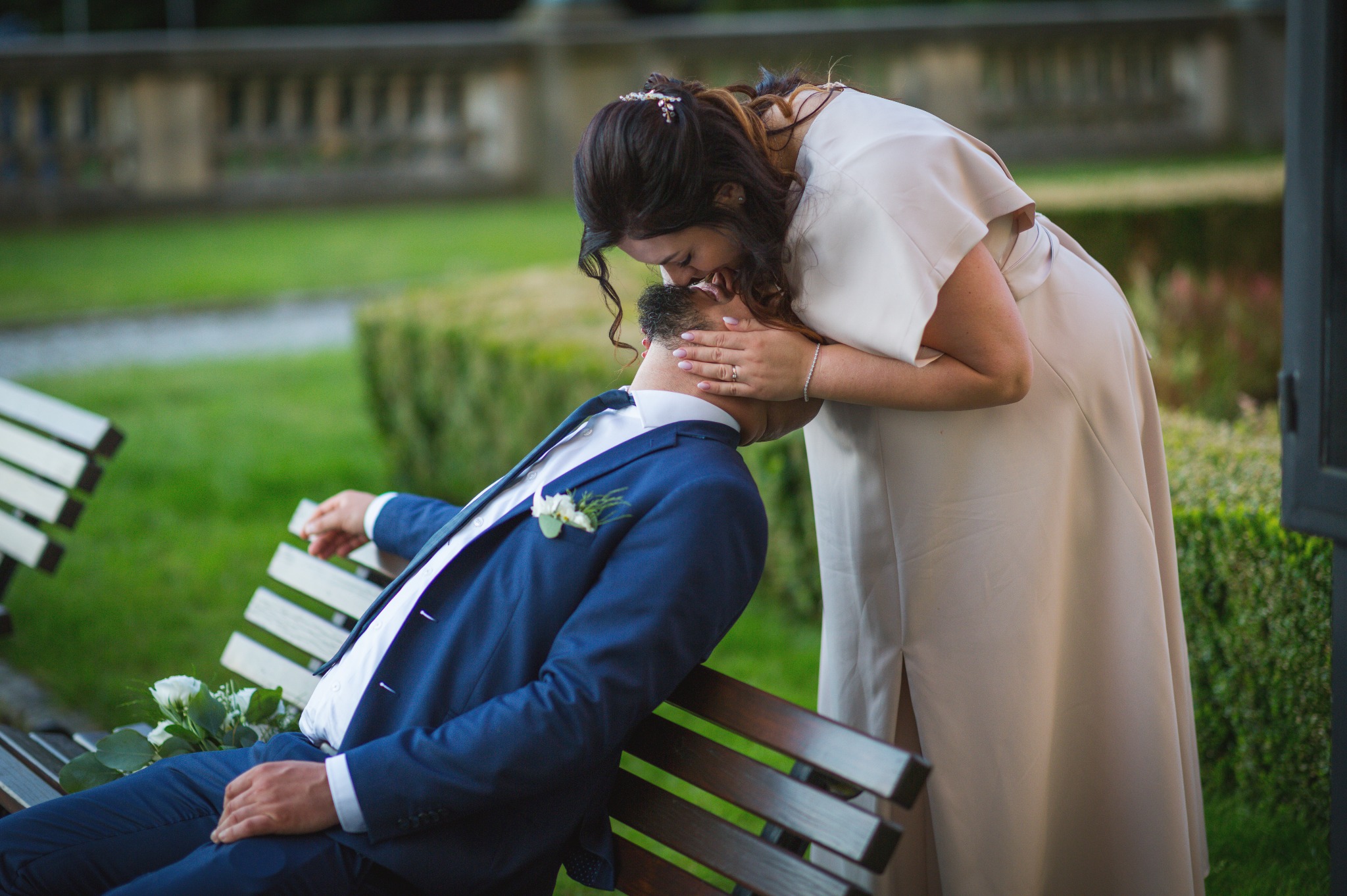 Panna młoda w beżowej sukni całuje siedzącego na ławce pana młodego w granatowym garniturze. W tle zieleń trawy i krzewów, romantyczna sceneria.