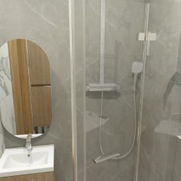 DYNAMIC ENGLISH LANGUAGE SERVICES SPÓŁKA Z OGRANICZONĄ ODPOWIEDZIALNOŚCIĄ - Nowoczesna łazienka z kabiną prysznicową, umywalką z szafką i owalnym lustrem. Ściany wyłożone płytkami imitującymi marmur. Widoczny dozownik na półce w kabinie.