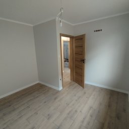LuxArt Usługi Remontowo-Budowlane Jakub Kusibab - Jasne wnętrze po remoncie w Przecławiu: drewnopodobna podłoga, świeżo pomalowane ściany na szaro, otwarte drewniane drzwi. Widoczne gniazdka elektryczne.