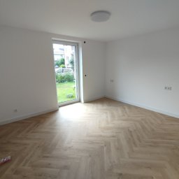 Jasne, puste wnętrze z parkietem ułożonym w jodełkę, świeżo pomalowane ściany na biało i widokiem na zieleń przez okno. Minimalistyczny design, gotowe do umeblowania.