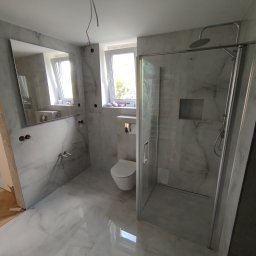 LuxArt Usługi Remontowo-Budowlane Jakub Kusibab - Wykończona łazienka w szarościach z kabiną prysznicową typu walk-in, lustrem i podwieszaną toaletą. Widoczne narzędzia i materiały budowlane.