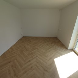 LuxArt Usługi Remontowo-Budowlane Jakub Kusibab - Jasne panele ułożone w jodełkę w pustym pokoju z białymi ścianami i oknem. Widoczne przygotowane punkty elektryczne na ścianie i narzędzie w rogu.