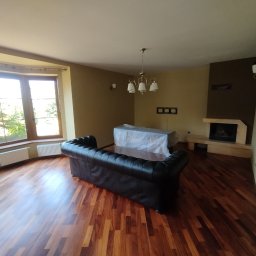 LuxArt Usługi Remontowo-Budowlane Jakub Kusibab - Salon z drewnianą podłogą, kominkiem i dużym oknem. Meble częściowo zakryte folią ochronną, sugerujące prace wykończeniowe lub przygotowanie do nich.