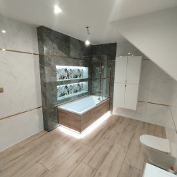 Nowoczesna łazienka po remoncie: marmurowe ściany z złotymi akcentami, podświetlana wanna obudowana drewnem, drewnopodobna podłoga, biała szafka i toaleta.