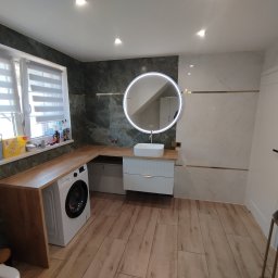 Nowoczesna łazienka z drewnianym blatem, pralką, okrągłym lustrem z podświetleniem LED i białymi płytkami imitującymi marmur z złotymi akcentami.