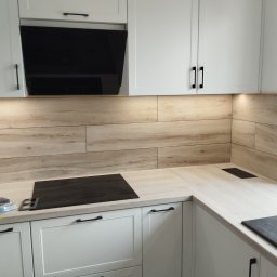 Jasna kuchnia z drewnopodobnymi płytkami na ścianie, czarnymi uchwytami i płytą indukcyjną. Widoczne detale wykończenia i oświetlenie podszafkowe.