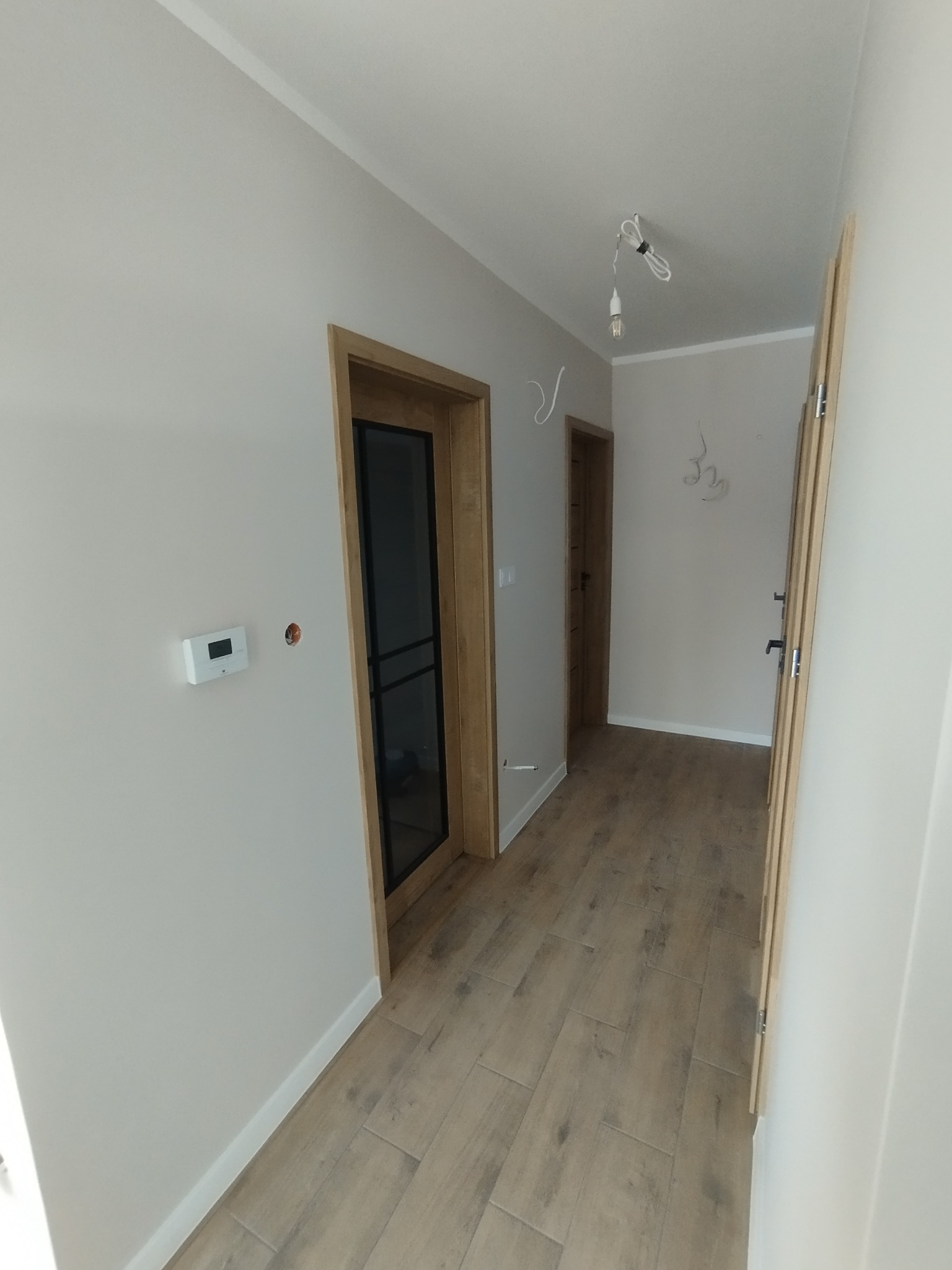 Jasne wnętrze korytarza po remoncie z drewnianą podłogą, dwoje drzwi z ościeżnicami i widocznym termostatem na ścianie. Minimalistyczny design.