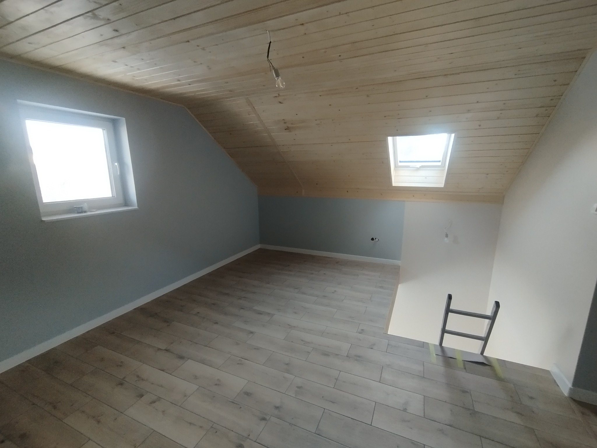 Jasne poddasze z drewnianą boazerią sufitową i panelami podłogowymi. Okno dachowe wpuszcza światło. Ściany pomalowane na szaro. Widoczna drabina.