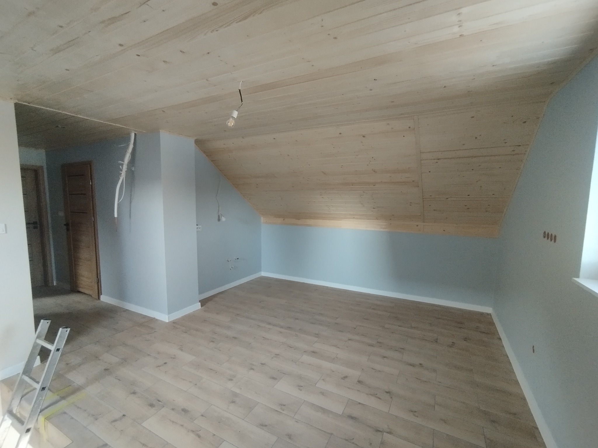 Jasne, wykończone poddasze z drewnianym sufitem i panelami podłogowymi. Ściany pomalowane na szaro, widoczna drabina malarska i wystające kable.
