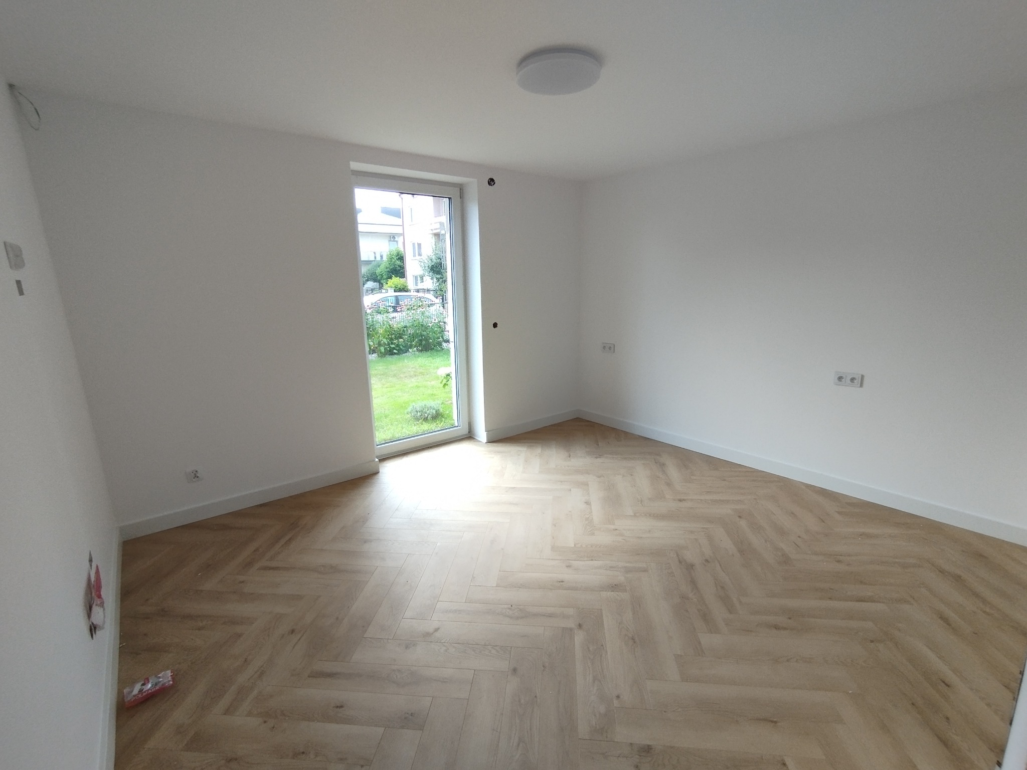 Jasne, puste wnętrze z parkietem ułożonym w jodełkę, świeżo pomalowane ściany na biało i widokiem na zieleń przez okno. Minimalistyczny design, gotowe do umeblowania.