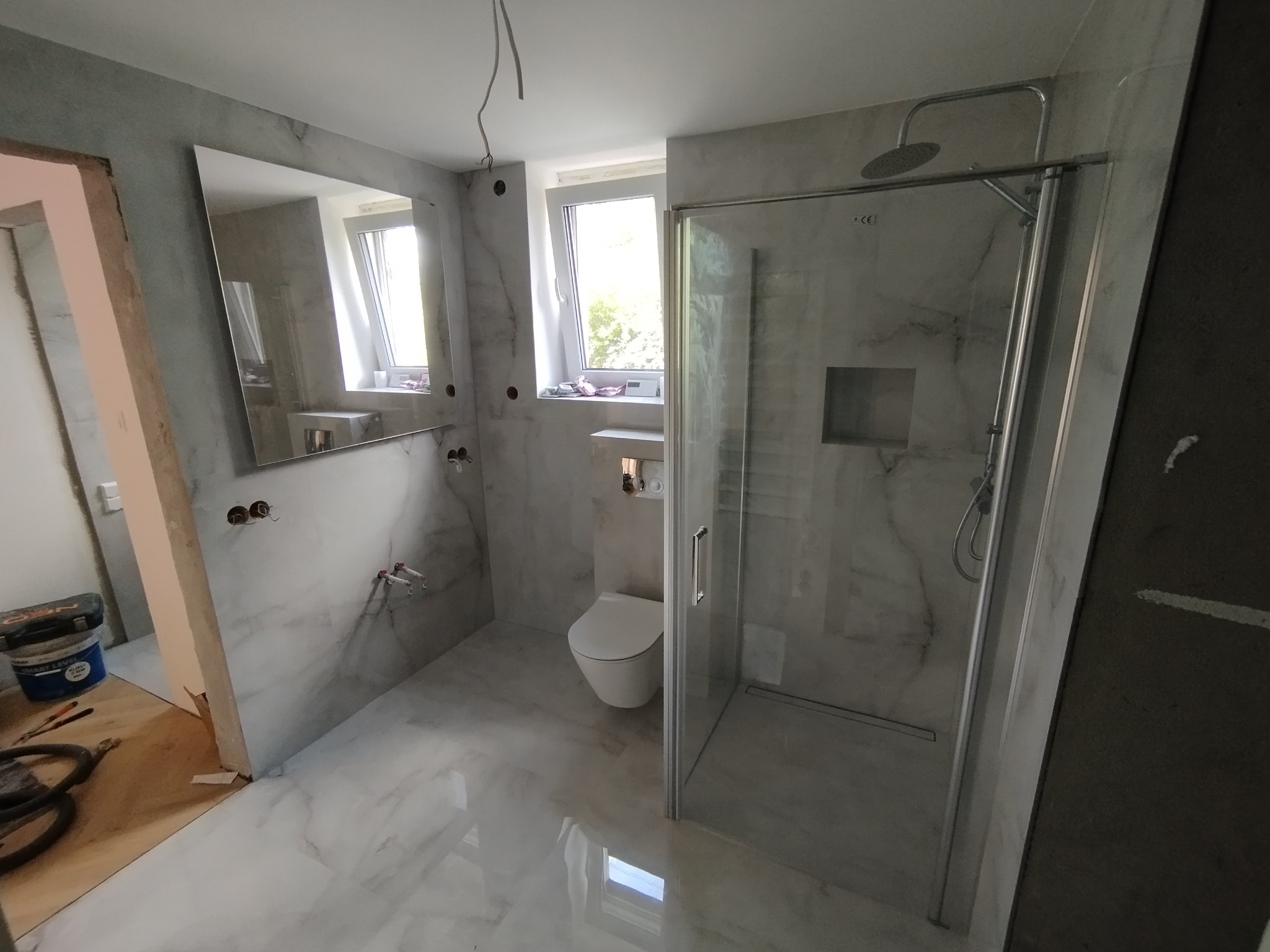 Wykończona łazienka w szarościach z kabiną prysznicową typu walk-in, lustrem i podwieszaną toaletą. Widoczne narzędzia i materiały budowlane.