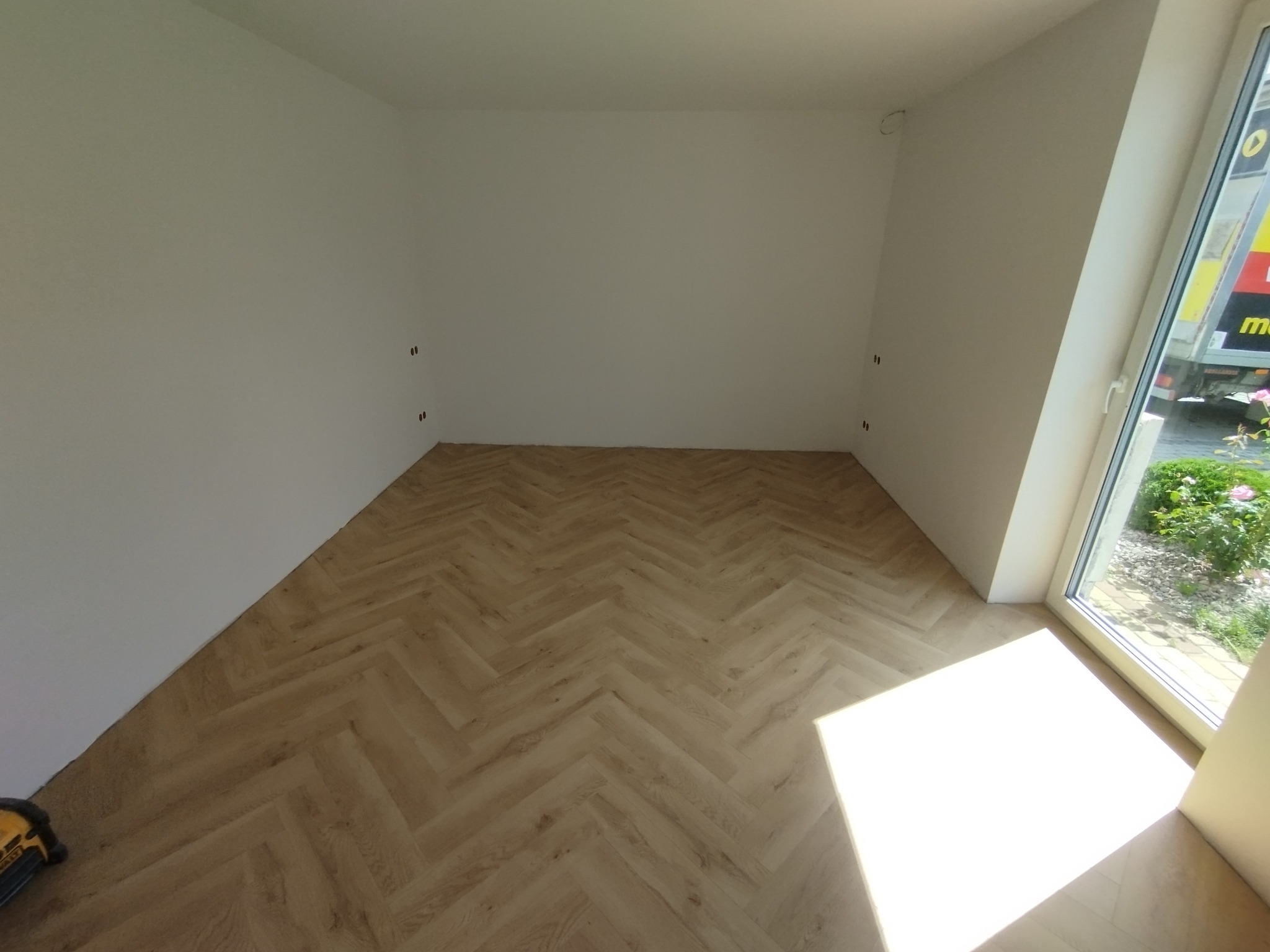 Jasne panele ułożone w jodełkę w pustym pokoju z białymi ścianami i oknem. Widoczne przygotowane punkty elektryczne na ścianie i narzędzie w rogu.