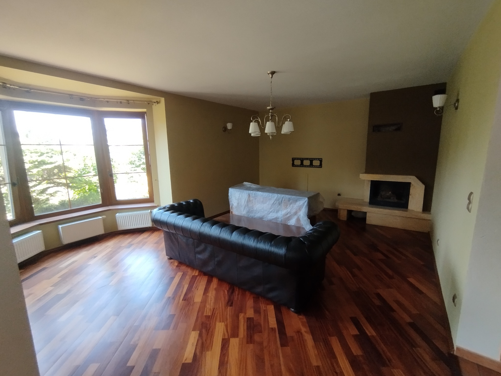 Salon z drewnianą podłogą, kominkiem i dużym oknem. Meble częściowo zakryte folią ochronną, sugerujące prace wykończeniowe lub przygotowanie do nich.