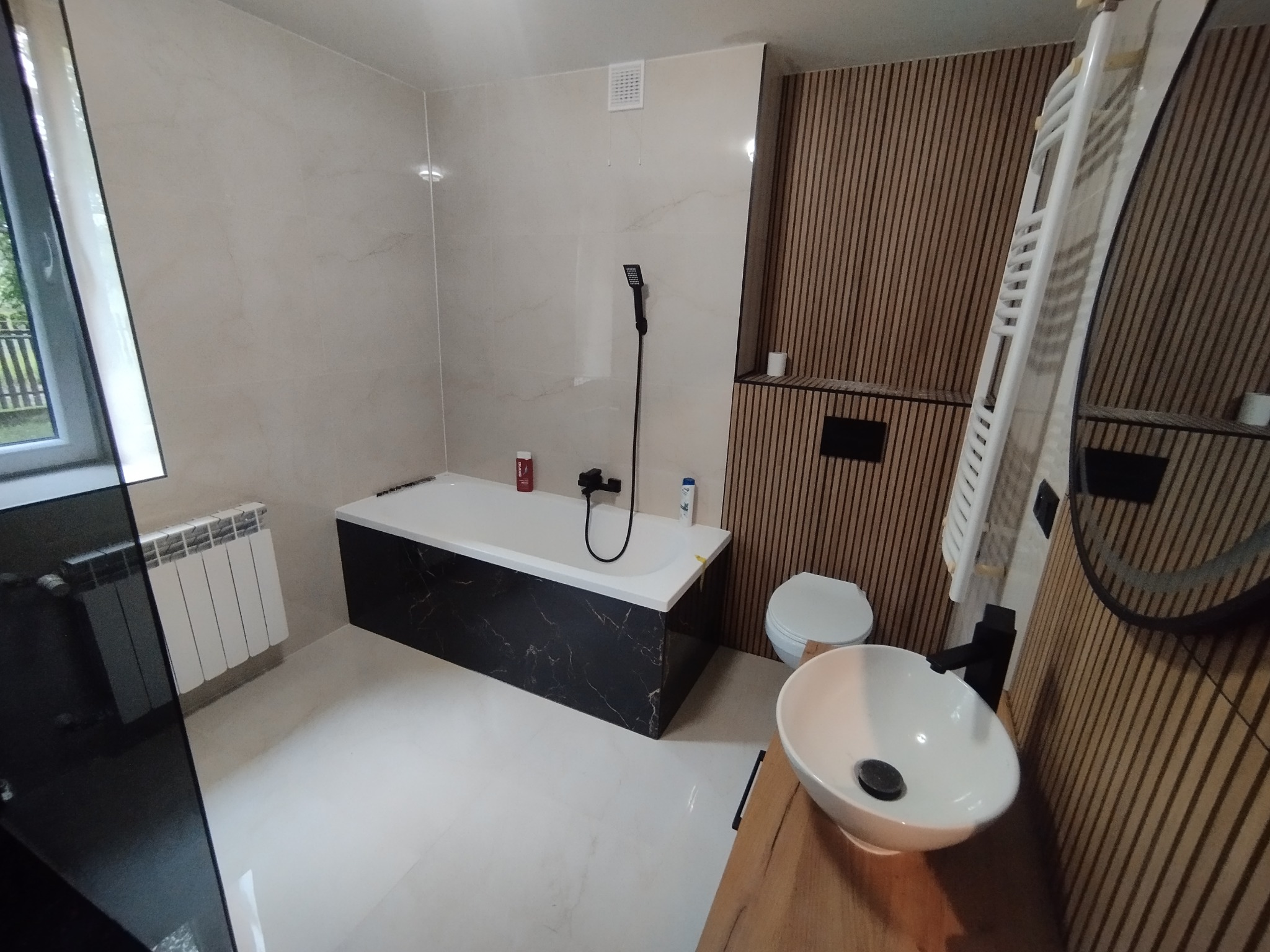 Nowoczesna łazienka po remoncie: beżowe płytki, wanna z czarnym marmurem, drewniana ściana z lamelami, biała umywalka nablatowa i grzejnik drabinkowy.