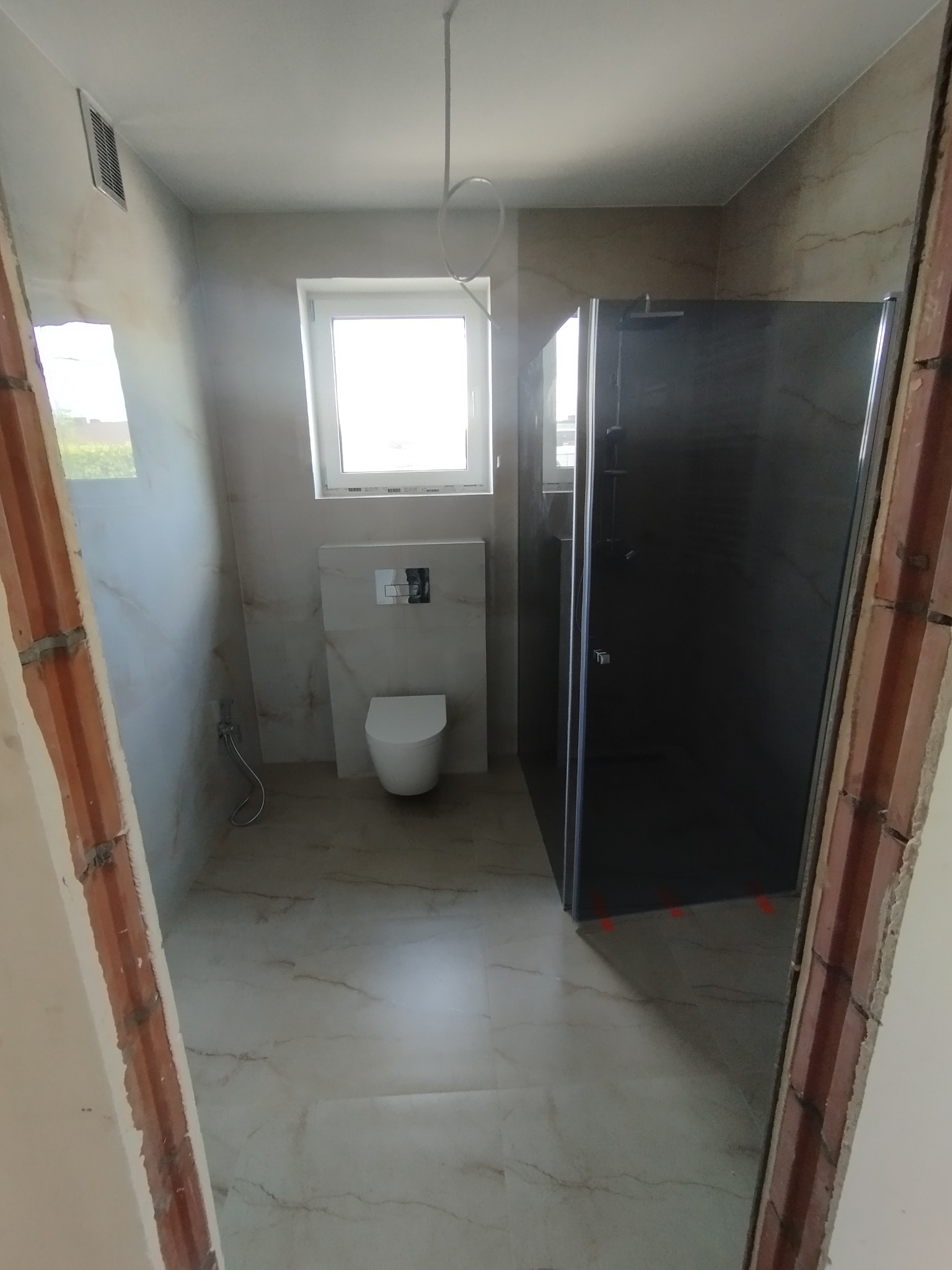 Nowoczesna łazienka w trakcie wykańczania: kabina prysznicowa z ciemnego szkła, podwieszana toaleta i marmurowe płytki. Widoczne surowe wykończenie ościeżnicy.