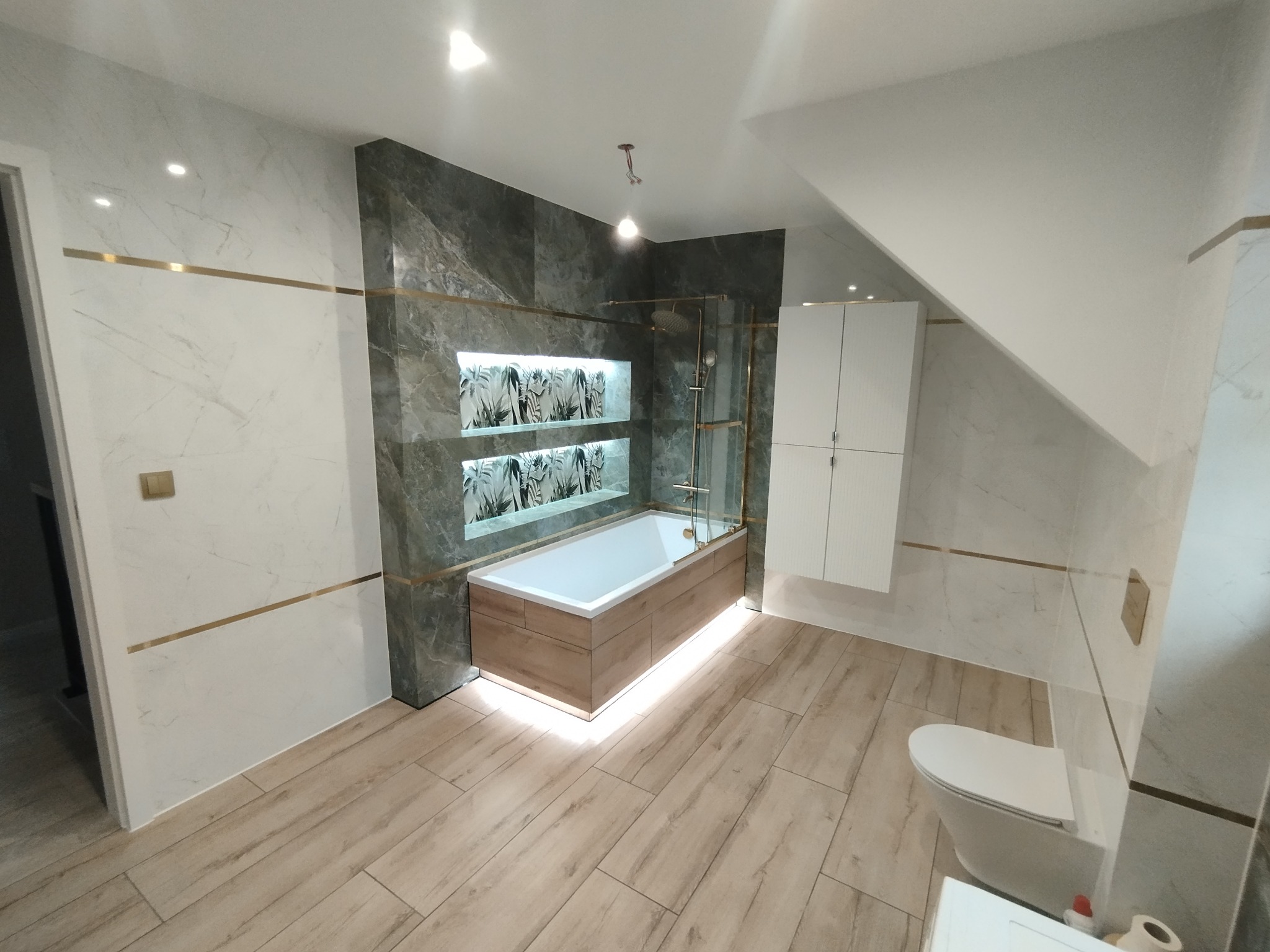 Nowoczesna łazienka po remoncie: marmurowe ściany z złotymi akcentami, podświetlana wanna obudowana drewnem, drewnopodobna podłoga, biała szafka i toaleta.