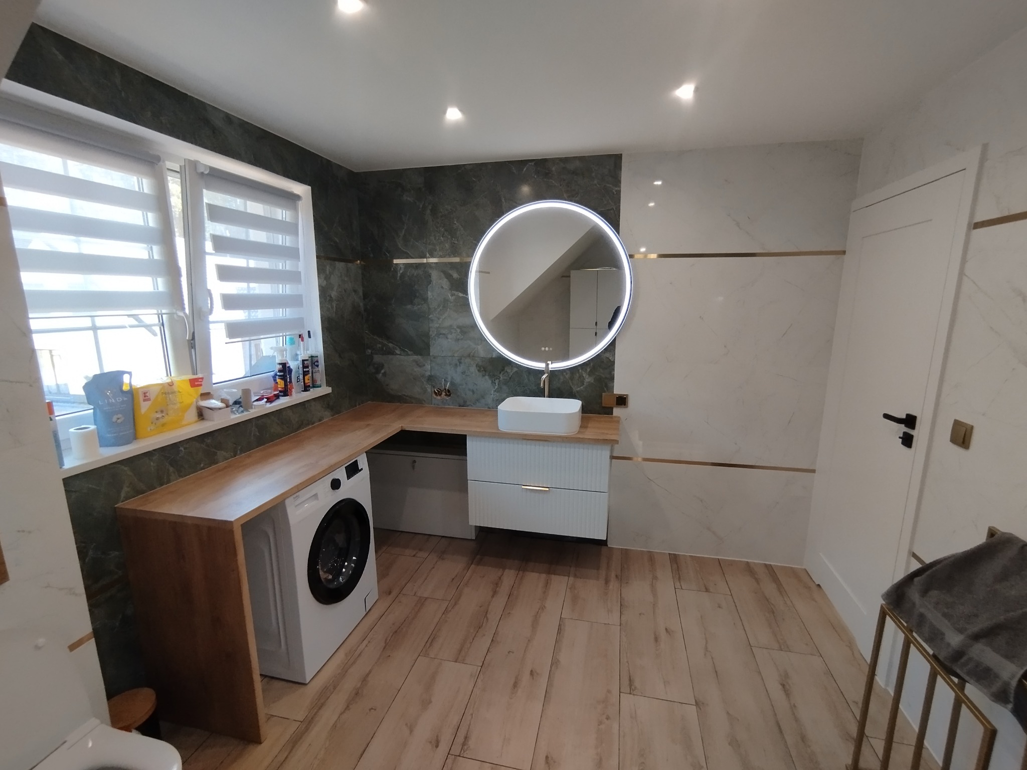 Nowoczesna łazienka z drewnianym blatem, pralką, okrągłym lustrem z podświetleniem LED i białymi płytkami imitującymi marmur z złotymi akcentami.