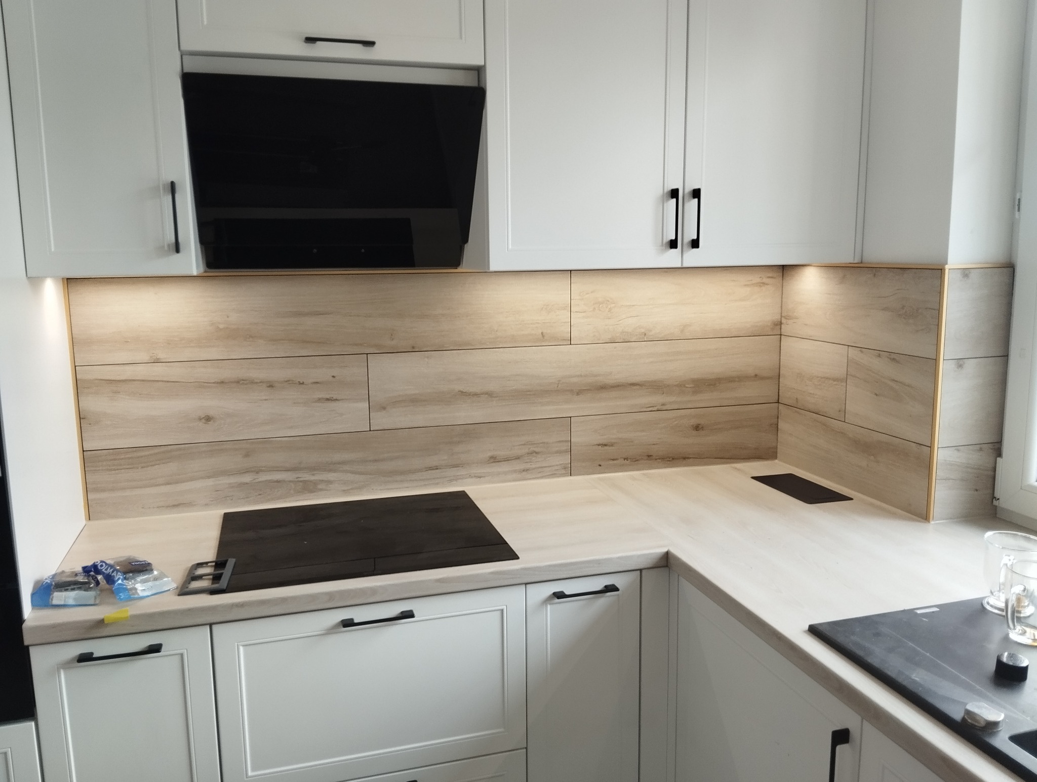 Jasna kuchnia z drewnopodobnymi płytkami na ścianie, czarnymi uchwytami i płytą indukcyjną. Widoczne detale wykończenia i oświetlenie podszafkowe.