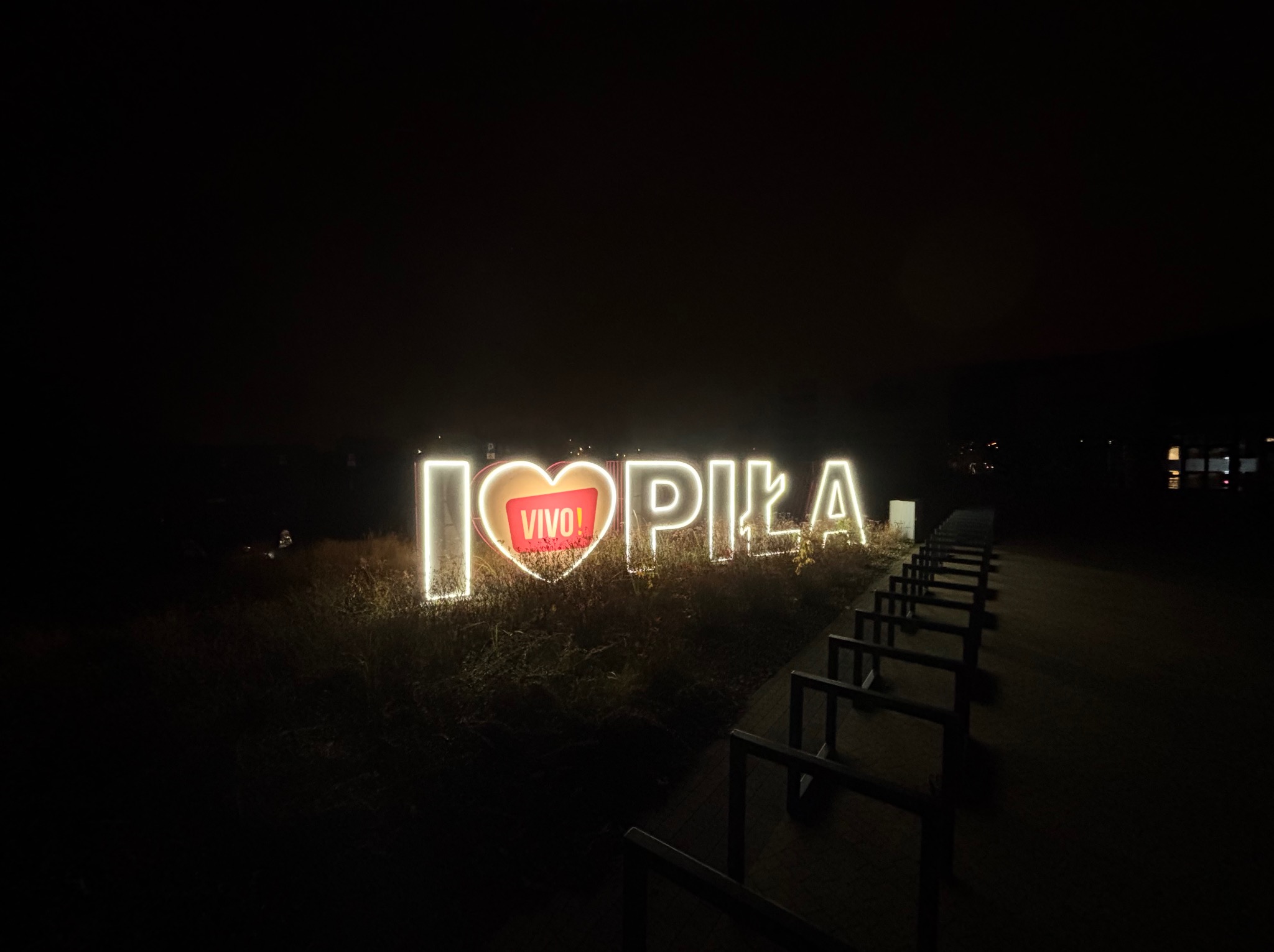 Iluminowany napis 'I love Piła' z czerwonym sercem z logo 'VIVO!' w środku, ustawiony na trawie w nocy, widoczne barierki wzdłuż ścieżki.