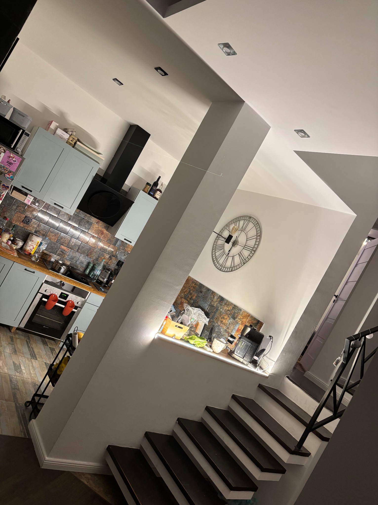 Nowoczesna kuchnia z szarymi szafkami, czarnym okapem i mozaiką nad blatem, widoczna z perspektywy schodów z czarną balustradą i dekoracyjnym zegarem na ścianie.