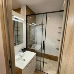 Nowoczesna łazienka z kabiną prysznicową typu walk-in z czarnymi profilami, białą umywalką nablatową z drewnianą szafką i lustrem z oświetleniem LED.