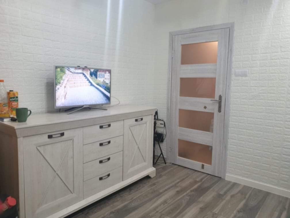 Salon w Besko z komodą w stylu rustykalnym, telewizorem i ścianami wyłożonymi dekoracyjną cegłą piankową. Widoczna również odnowiona, przeszklona wewnętrzna stolarka drzwiowa.